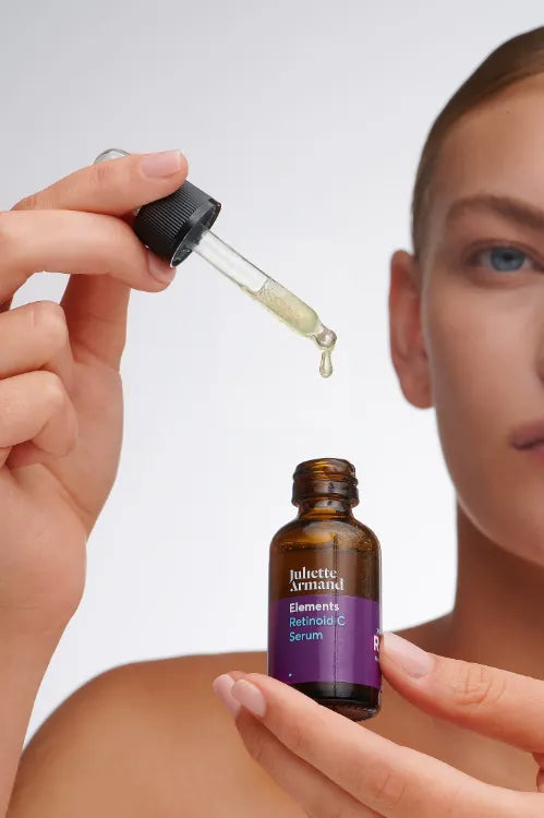 Close-up van een vrouw die het Juliette Armand Elements Retinoid C Serum gebruikt. Ze houdt het bruine flesje vast terwijl een pipet met een druppel serum boven de opening hangt.
